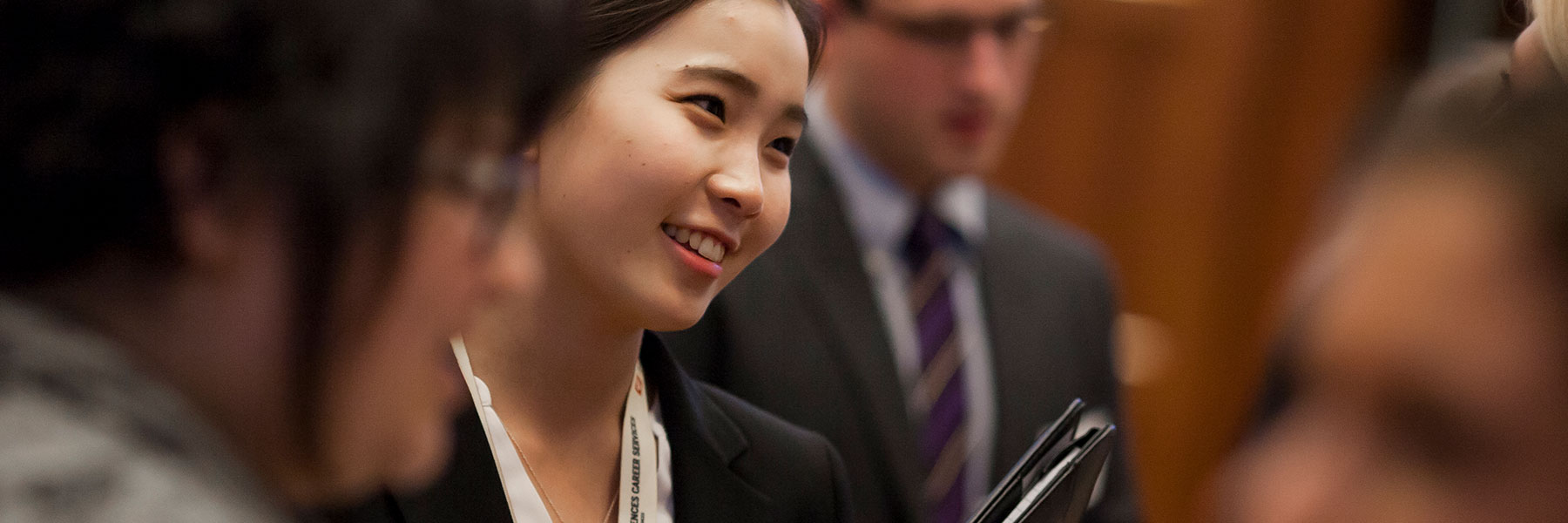 Young woman in formal businesswear holds a folder as she speaks with people at a career event.