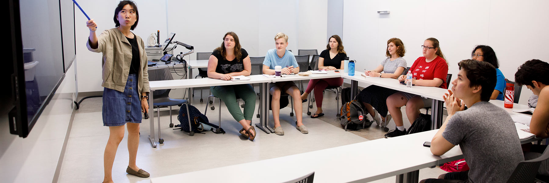 An instructor points to a wall-mounted screen as students seated in the classroom listen and watch.