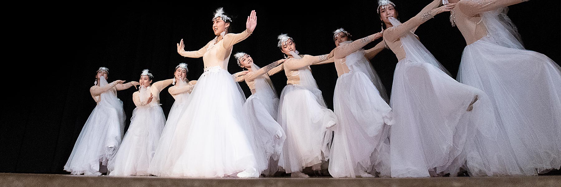 A group of women in white dresses form a line dancing with one woman in front with her arms open wide.