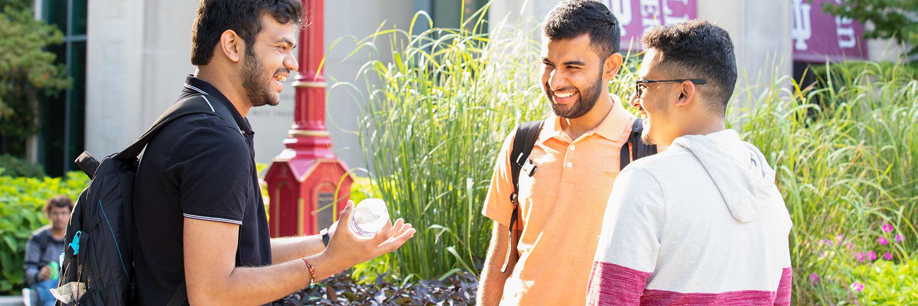 Three male students talk outside on campus with plants and flowers in the background.