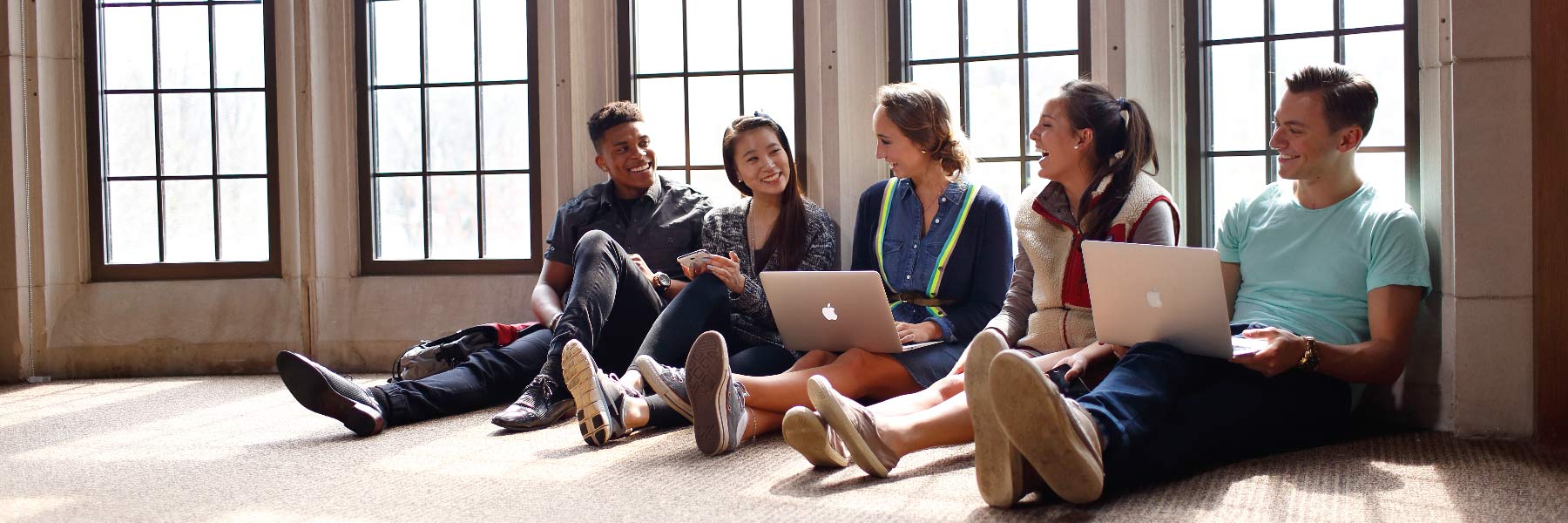 Five students sit on the floor with their backs against windows along the wall.